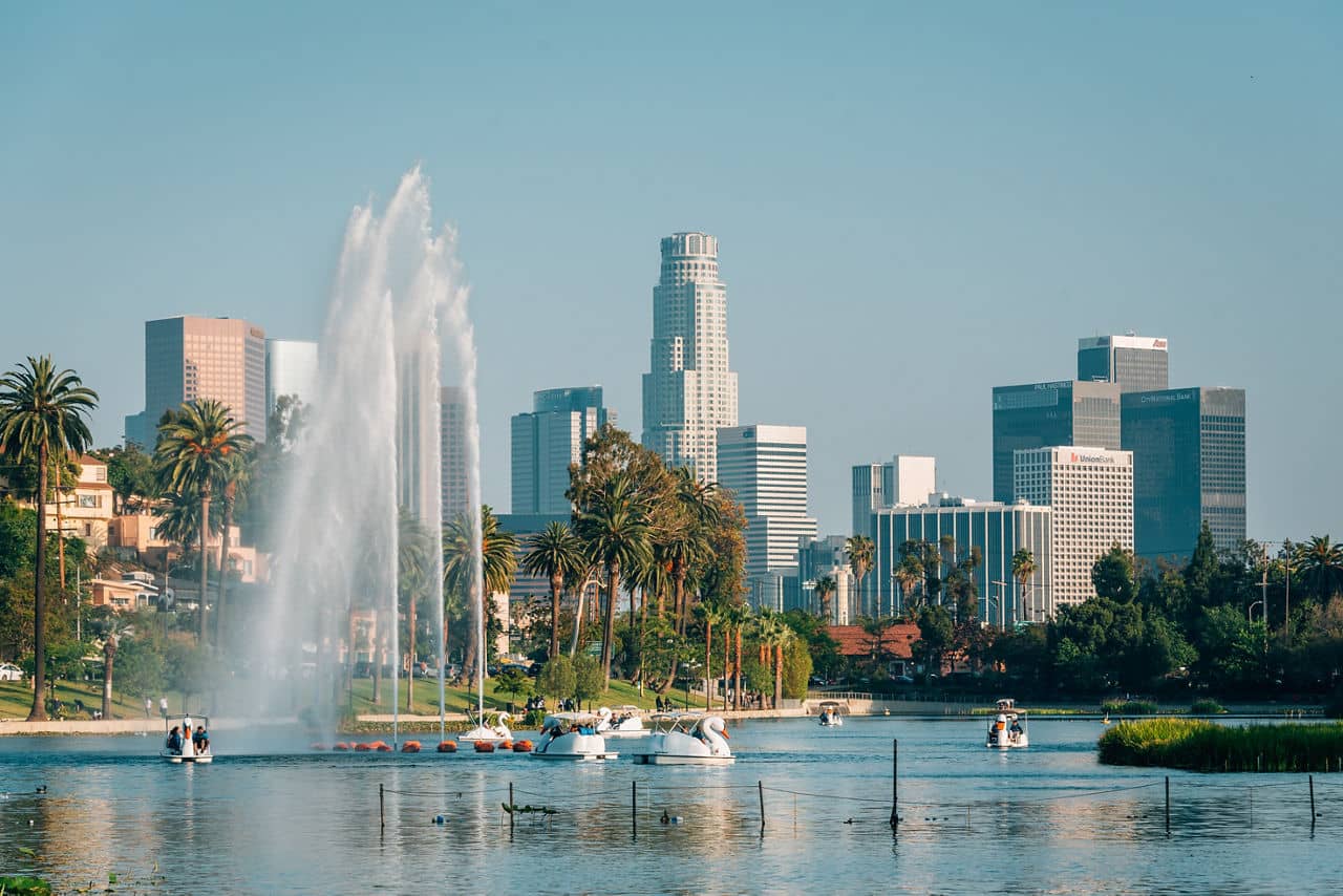 A scenic view of Echo Park, California, featuring a lake with paddle boats, tall fountains, and the Los Angeles skyline in the background.